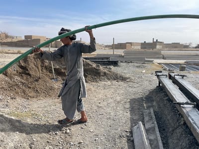 An outdoor scene in Nakhonay, Afghanistan, depicts a young male at a construction site under a clear, bright blue sky. The individual, dressed in a grey traditional garment and dark sandals, wears a dark turban and holds a long, green pipe with both hands above his head, creating an arc. He is positioned left of center, looking towards the right and slightly downwards. To his left, a large mound of reddish-brown earth contains two upright shovels. The ground is dry, light-colored soil mixed with gravel. To his right, two long, dark concrete molds or beams rest on low supports, with additional stacks of pre-cast concrete slabs and piles of rebar visible further back. In the background, a dirt road runs horizontally behind a sparse fence. Beyond the road, several single-story buildings constructed of light-colored brick or mud brick, some appearing incomplete or in disrepair, are visible. One building exhibits a prominent chimney-like structure.