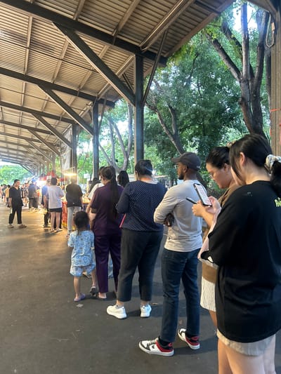 The image depicts a street scene at dusk in Tha Khlong Town Municipality, Thailand. A covered walkway with a corrugated metal roof supported by metal beams stretches into the distance, illuminating a food stall area with overhead lights. A line of people, varying in age and gender, are queuing. In the foreground, a woman in a black shirt and shorts faces away from the camera, while to her right, a young girl in a light blue patterned outfit stands with her back to the viewer. Further back, several individuals are visible in the queue, some looking at their phones. The ground is paved with asphalt. Trees with green foliage are visible behind the walkway, suggesting an outdoor market setting. The overall atmosphere appears casual and public, likely a common gathering place for food or shopping.