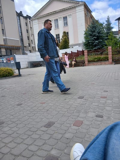 The image depicts a paved walkway in Truskavets, Ukraine, likely near a building with classical architectural elements, possibly a theater or cultural center, indicated by the columns and pediment visible in the background. A man in a denim jacket and jeans is walking towards the right of the frame, with his left side to the camera. He is wearing dark sneakers. Behind him, a woman with dark hair and glasses is walking, also towards the right, and a person in pink pajamas is holding a phone and looking down. Further back, another woman is seated on a bench. The walkway is paved with interlocking grey bricks. There are trees, a fence, and a trash can visible in the scene. The sky is overcast with some blue patches.