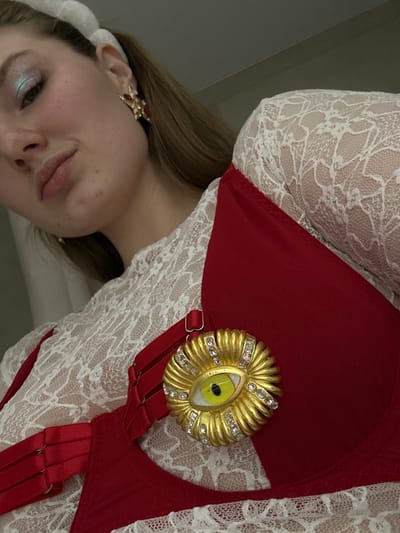 A woman with fair skin and light brown hair is visible from the shoulders up. She wears a white lace top under a red garment resembling a bra or corset with several straps. A gold brooch shaped like an eye, adorned with small gems, is pinned to the red fabric. Her makeup includes light blue eyeshadow. A decorative earring is visible on her left ear.