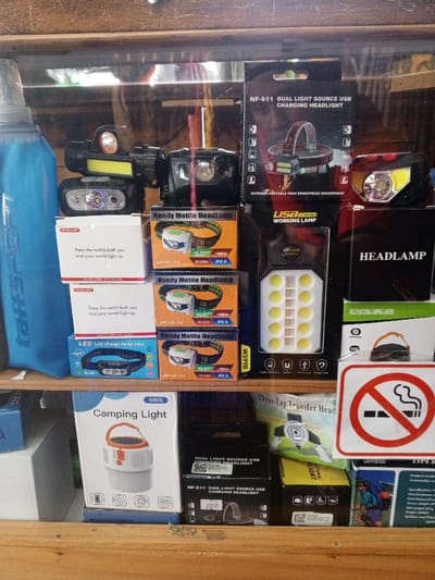 The image displays a retail shelf or display case filled with various headlamps and portable lighting devices, likely in a store or market in Baza, Myanmar. The scene is dominated by product packaging and unpackaged light units, indicating items for sale.
On the far left, a tall, blue "taffSPORT" branded water bottle stands next to a stack of white boxes. These boxes, labeled "HEADLAMP," feature the slogan "Press the switch, both you and your world light up." Below them, a blue box advertises an "LED Led charge headlamp KL-05," and a white box for a "5805 Camping Light" is visible.
Multiple orange boxes for "Mandy Motile Headlamp" are stacked in the center-left, with details like "LED," "ART NO. T16," "50 000%," and "APX 6" printed on them. Above these boxes, several headlamps are displayed out of their packaging, showcasing different designs, including some with multiple light sources and head straps.
Towards the right, black boxes for "NF-611 DUAL LIGHT SOURCE USB CHARGING HEADLIGHT" are visible, highlighting features like "OUTDOOR PORTABLE HIGH BRIGHTNESS WATERPROOF." One box shows "85000," which may be a price. A "USB WORKING LAMP W599B" is displayed in its packaging, featuring a panel of small LED lights. Other black boxes are labeled "HEADLAMP" and "MULTIFUNCTIONAL CAMPING LANTERN." Further down, a green package for a "Three-Leg Transfer Head" and a box from "KOVAR TYPE S" showing a hiker graphic are present.
In the bottom right corner, a prominent "No Smoking" sign—a white circle with a red border and a diagonal line over a black cigarette—is affixed to the shelf. The display is somewhat cluttered but organized, resting on a wooden surface, with reflections suggesting a glass-fronted case. The objects are under ambient indoor lighting. No specific activity, time of day, or weather is depicted beyond the retail setting.
