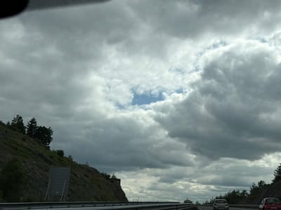 This is a landscape photograph taken from a moving vehicle on a highway. The sky is overcast with heavy, dark clouds, but there is a patch of blue sky visible in the upper center. To the left, a steep, grassy hillside is covered with trees, and a metal guardrail runs along the bottom of the image. Two cars are visible on the highway ahead, suggesting the vehicle the photo is taken from is also on the highway. The scene appears to be on the outskirts of Staro Selo, Bulgaria, given the characteristic landscape of the region. The overall mood of the image is somewhat somber due to the cloudy sky.