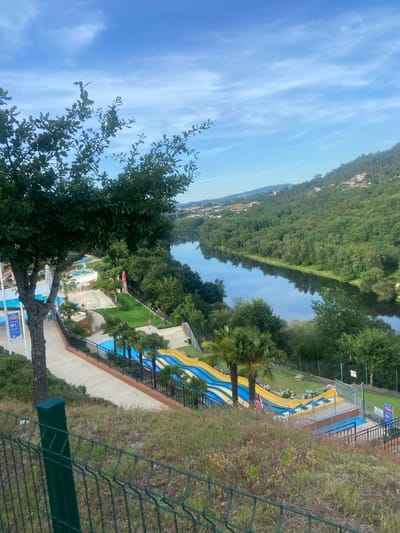 The image shows an aerial view of a water park with several slides, pools, and lounging areas. The park is situated on a hillside overlooking a wide river, the Tâmega River. Lush green trees and vegetation cover the surrounding hillsides, with houses and buildings visible in the distance on the slopes. The sky is a clear blue with scattered white clouds. In the foreground, a large tree with dark green leaves stands to the left, and a green metal fence runs along the bottom of the frame. The water slides are colorful, with blue and yellow stripes. Palm trees are scattered throughout the park. The scene suggests a sunny day at a recreational facility. The location is identified as Amarante, Portugal.