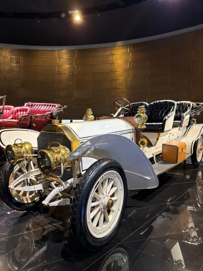 The image shows a white antique car with brass accents, parked on a highly reflective black floor. The car has large, spoked wheels, prominent brass headlamps, and a leather-upholstered interior. A wooden chest is visible on the side of the car. In the background, a curved wall with a textured golden surface is illuminated by overhead lights. Another antique car with red leather seating is partially visible to the left. The scene appears to be an indoor exhibition or museum, likely the Mercedes-Benz Museum in Stuttgart, Germany, given the context provided. There are no people visible in the image. The lighting is dramatic, with spotlights creating highlights and reflections on the polished surfaces.