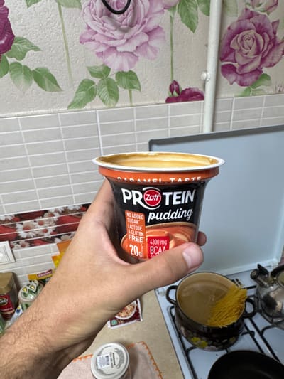 A human hand, appearing to be an adult male's right hand, holds an open container of Zott Protein Pudding, Caramel Taste. The pudding container is black with an orange band and white lid rim, displaying "NO ADDED SUGAR," "LACTOSE & GLUTEN FREE," "20g PROTEIN," and "4300 mg BCAA" text. The caramel-colored pudding is visible within the container. The hand and forearm exhibit light skin tone and moderate hair.
The scene is an indoor kitchen environment in Ciorescu, Moldova. The background features a wall adorned with two distinct wallpaper patterns: an upper section with large pink and purple roses and green leaves on a light background, and a lower section (partially visible to the left) with smaller, daisy-like floral patterns in orange, white, and brown. Between these wallpaper sections, a horizontal strip of light gray, rectangular tiled backsplash is present. A white vertical pipe is affixed to the wall on the right.
On a white countertop to the lower right, a dark-colored ceramic or enamel pot with floral designs sits on a stovetop burner, containing water and partially submerged thin, yellow strands of pasta or noodles. A metallic kettle is partially visible to the right of the pot. To the lower left, on the countertop, a small white plastic container with a barcode label, an orange and yellow snack packet (partially visible, possibly "Galeo"), and other indistinct packaged items are present.