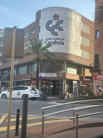 The image shows the exterior of the "Centre Comercial Carabela" shopping center in Lloret de Mar, Spain. The building features a large, circular facade with the shopping center's name and logo prominently displayed. The logo consists of stylized letters "CCC" and a red sailing ship. The building is made of brick and has several floors with windows and balconies. In front of the shopping center, there are palm trees, shops, and pedestrians. A white car with "WATER WORLD" branding is parked on the street. The scene is captured on a sunny day, with clear blue skies.