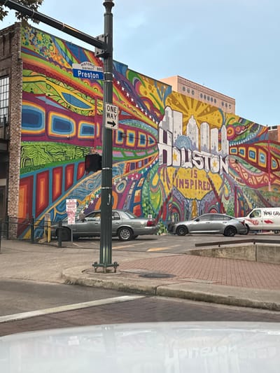 A vibrant mural adorns a building, depicting the city of Houston with colorful geometric designs. The word "Houston" is prominently displayed above the skyline. A street sign indicates "Preston" in the historic district. A "One Way" sign is also visible. Cars are parked along the curb next to a brick sidewalk. A traffic light pole stands on the corner.