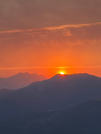The image captures a dramatic sunset over a mountainous landscape, oriented vertically. The sun, a bright orb of golden light, is partially obscured by the silhouette of a large, dark mountain range on the right side of the frame. The sky is ablaze with warm hues of orange and yellow, indicating either sunrise or sunset. Wispy clouds, tinged with the same warm colors, are visible on the left, adding texture and depth to the sky. The mountain range appears rugged and imposing, with subtle variations in shade suggesting depth and distance. The overall scene evokes a sense of awe and natural beauty. The location context suggests this scene is likely in or near San Juan de Plan, Spain, a region known for its mountainous terrain. The time of day is clearly either dawn or dusk. No people or objects are visible, focusing the viewer's attention entirely on the natural elements of the landscape. The emotion conveyed is one of peacefulness and the grandeur of nature. There is no visible text in the image.