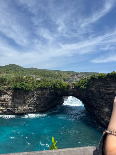 The image displays an expansive, elevated view of Broken Beach (Pasih Uug), a natural rock arch formation located near Sakti village on Nusa Penida island, Klungkung Regency, Indonesia.

In the immediate foreground, the lower right quadrant reveals a portion of a human arm and shoulder, clad in dark fabric, and a wrist adorned with a leopard-print bracelet. A dark grey ledge or railing extends across the bottom, with a small plant featuring bright green leaves sprouting from it. The central focus is a massive natural rock arch, predominantly dark grey and brown, spanning a channel of brilliant turquoise ocean water. White foamy waves are visible crashing into and through the arch. The rugged cliffs forming the arch are partially covered with green vegetation, including bushes and small trees. Beyond the arch, the ocean water appears a deeper blue with visible surface ripples. In the background, rolling green hills, densely covered in foliage, extend towards the horizon under a bright blue sky scattered with wispy white clouds. A small, light-colored structure is discernible on one of the distant hills. On the cliff edge above the right side of the arch, at least one human figure is visible, standing and observing the scenic vista.