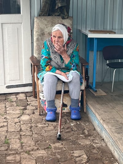 An elderly woman, wearing a white headscarf and a floral-patterned blouse with a grey embroidered vest, sits on a chair. She has her hand covering her mouth and appears to be either coughing or speaking. The woman is wearing grey pants and blue boots. A cane with a black handle rests beside her, its tip on the stone ground. The setting appears to be outdoors, possibly a porch or patio, with a wooden floor and a brick or stone wall in the background. To the left, there's a white door and part of a building structure with corrugated siding. A blue shelf unit with a dark chair is visible behind the woman. A brown bottle lies on a shelf within the blue structure. The overall scene suggests a casual, perhaps domestic, outdoor setting. The lighting and shadows imply it is daytime.