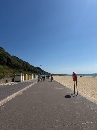 The image captures a sunny day on the promenade and beach in Bournemouth, United Kingdom. A paved walkway runs along the left side of the image, bordered by a road and a row of colorful beach huts. The beach huts are varied in color, with white, yellow, and green being prominent. Behind the beach huts, a green, treed hillside rises steeply.
On the promenade, several people are walking, appearing relaxed and enjoying the weather. They are dressed in casual attire, suggesting a typical day at the seaside. To the right of the promenade is a wide expanse of golden sand, the beach. In the foreground of the beach, a red and white life buoy on a stand is prominently positioned. Further down the beach, other beachgoers can be seen, some appearing to be sitting or standing. The sea is visible in the distance, a calm blue under a clear, bright sky. The overall scene conveys a peaceful and pleasant atmosphere of a popular seaside resort.