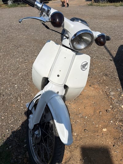 The primary subject of the image is a white Honda motorcycle or scooter, viewed from an elevated, oblique angle, parked outdoors. The vehicle features a large, circular chrome-rimmed headlight with a ribbed lens and two smaller, dark spherical indicator lights above it. The handlebars are visible on the left, showing a chrome lever and a black grip. A distinct "HONDA" logo, featuring stylized wings within an oval, is prominently displayed on the white bodywork to the right of the handlebars. The front wheel, with chrome spokes and a black tire, is partially visible, covered by a white mudguard.
The motorcycle is situated on a rough surface composed of gravel, small stones, and dry soil, with some patches of grass visible towards the bottom right. Strong, dark shadows are cast on the ground to the right, indicating bright, sunny daylight conditions. In the far left background, a portion of a person's feet and legs, wearing sandals, can be seen. The vehicle appears to be stationary, with no specific activity occurring. The scene depicts a casual outdoor setting on a dry, unpaved surface. The visible text is the "HONDA" brand name on the vehicle's logo.