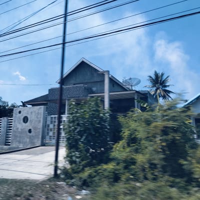 The image captures a residential scene in Keudelapang, Indonesia, taken from a moving vehicle, evidenced by the motion blur in the foreground. The prominent subject is a modern-style house with a grey, angular roof and concrete walls, situated under a clear blue sky with sparse white clouds. Multiple dark utility wires stretch across the upper portion of the frame.

The house features a covered patio or carport area and a satellite dish mounted on its roof. Lush green trees and bushes frame the house, with a tall palm tree distinctly visible behind it, suggesting a tropical environment. To the left of the house, a grey concrete fence or wall with distinct circular cutouts and a gate provides a boundary. A concrete driveway or access road leads towards the house from the foreground. A dark utility pole is partially visible on the right side of the driveway. The overall setting depicts a sunny daytime scene in a seemingly quiet, residential area. No specific activity or visible text is present.