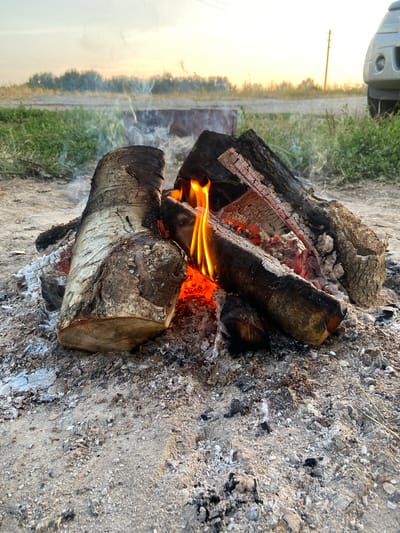 A campfire is burning outdoors on a sandy ground. The fire is made of several logs, some of which are partially burnt, and embers glow with orange and red light. Smoke rises from the fire. In the background, there is green grass and a dirt road. A vehicle is visible on the right side of the image, and the sky is a soft yellow and white, suggesting it is either sunrise or sunset. The location is identified as Peshcherka, Russia.