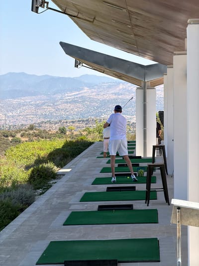 In Tsada, Cyprus, at a golf driving range, a person wearing a white polo shirt, white shorts, and a blue baseball cap stands on a green mat, mid-swing with a golf club. To their left, another person in pink shorts and a white top is visible. The driving range is characterized by a tiled walkway, multiple green practice mats, and a basket of golf balls. White pillars support a large, angled wooden roof structure. In the background, a vast, dry, mountainous landscape stretches under a clear blue sky. The scene is illuminated by bright, natural daylight.