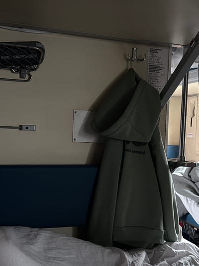 The image displays the interior of a train compartment, located within a carriage associated with Kargat, Russia. The scene is static and dimly lit. On the upper left, a dark-colored overhead luggage rack with a mesh base, marked "SMARTFORM," is affixed to a cream-colored wall. Below it, a small, horizontal rectangular fixture is mounted. Centrally positioned, a triple silver-toned metal hook suspends a dark green hooded sweatshirt, partially revealing the text "look around" on its front. Adjacent to the hook on the right, a white laminated sign displays instructions in Russian text and Braille regarding the operation of a shelf or bunk, specifying how to raise it until fixators click and how to lower it. Below the sign, a white square panel secured by four screws is visible. The lower section of the image features a dark blue upholstered element, possibly a backrest, over which rumpled white bedding lies on a bunk. To the far right, a vertical metallic bar and a glimpse of a door or window frame are present, revealing another blue surface beyond. No individuals are present in the scene.