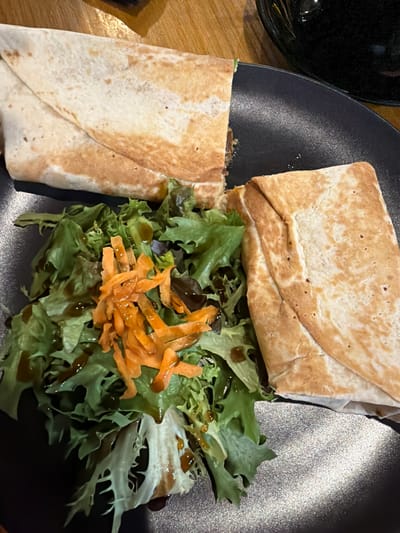 A close-up, overhead shot shows a black plate with two halves of a gyro and a side salad. The gyro, made with a wheat tortilla, is cut in half and appears to be filled with meat and some greens. The salad consists of mixed greens, shredded carrots, and a dark dressing drizzled over it. The plate is on a wooden surface, and a dark, reflective object is visible in the upper right corner of the image. The scene is indoors, likely a restaurant. The location is in the city of Epsom and Ewell, United Kingdom.
