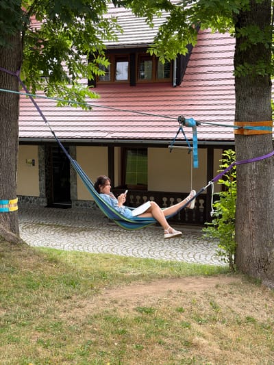 A woman is relaxing in a hammock strung between two trees in front of a building with a red tiled roof. The hammock is blue and green. The woman is wearing a denim jacket and shorts and is holding a phone in her left hand and a book in her right. She is barefoot with white sandals on her feet. The ground in front of the building is paved with cobblestones. There are green trees and grass in the foreground. The building appears to be a house or guesthouse. The scene is consistent with a leisure area in Świeradów-Zdrój, Poland, a resort town known for its spas and natural beauty.