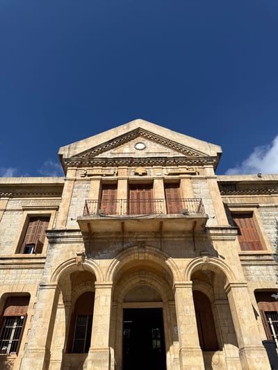 The image shows a low-angle, exterior shot of the historic Serail of Jezzine, Lebanon, under a clear, bright blue sky with a few wisps of white clouds. The building is constructed of light-colored stone blocks. Its architectural style features a triangular pediment adorned with decorative molding and a circular emblem. Below the pediment, there are two recessed windows with wooden shutters. A prominent feature is a weathered, ornate metal balcony supported by brackets, situated above a central arched entrance. The entrance consists of three large arches, with the central one leading into a dark interior. Flanking the central arches are two windows with arched tops, also featuring wooden shutters and iron grates on the lower left window. Shadows cast by the building's architectural elements create a play of light and dark across the facade. The overall impression is one of aged grandeur and historical significance.