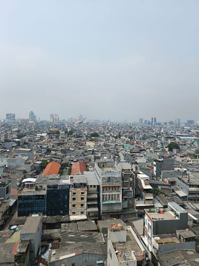 The image displays an expansive aerial view of a densely populated urban area within the Special Capital Region of Jakarta, Indonesia. The scene is observed under a light gray, hazy sky, indicative of an overcast day or atmospheric conditions common to large cities.
The foreground and midground are dominated by a vast expanse of tightly packed, low-to-mid-rise buildings, ranging from one to approximately eight stories. These structures exhibit diverse architectural styles and materials, with many featuring grey, corrugated metal, or dark-tiled roofs. Prominently visible are a multi-story building with dark, reflective glass panels, an adjacent light-colored building with multiple windows, and a distinct white/light gray structure characterized by vertical window elements. Numerous rooftops are equipped with utilitarian objects such as white and orange water tanks and various satellite dishes. Scattered reddish-brown tiled roofs suggest older or specific residential structures within the dense fabric. Due to the elevated vantage point and distance, no individual people or specific actions are discernible.
In the far background, the cityscape extends to the horizon, marked by a skyline of numerous taller, modern high-rise buildings and skyscrapers, presenting a stark contrast to the older, more densely packed structures in the closer view. Minimal vegetation is visible, limited to isolated trees interspersed among the buildings. The environment conveys a sense of extensive urban sprawl and high-density development, typical of Jakarta's mature districts transitioning towards modern commercial centers.