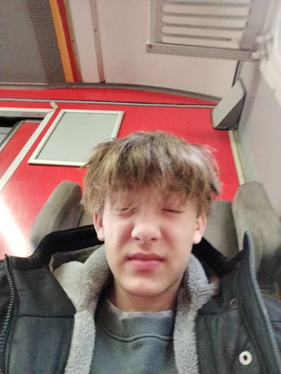 A young person with messy brown hair and eyes tightly shut sits in a seat, possibly on public transportation. They wear a grey sweater with a fluffy collar and a dark jacket. The interior features red walls, a white frame around a rectangular space, and overhead compartments. The person appears tired or uncomfortable.
