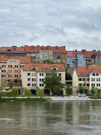 The image depicts a view of the Drava River embankment in Maribor, Slovenia, with a row of residential buildings lining the opposite bank. The sky is overcast with gray clouds. The buildings are multi-story structures with red tiled roofs, typical of European architecture. They vary in color, including beige, light brown, blue, and white. Several windows and balconies are visible on the facades.
Along the riverbank in the foreground, there is a paved walkway with some greenery, including weeping willow trees. There are a few people strolling on the embankment and some outdoor seating areas. A white car is parked on the walkway. The river itself is wide and reflects the overcast sky and the buildings. The water surface shows ripples and currents, indicating a moderate flow. The overall scene conveys a peaceful urban riverside setting under a cloudy sky.