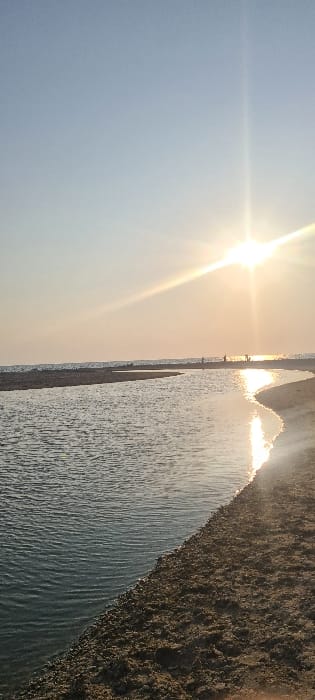 The image shows a view of the beach in Mikhmoret, Israel (latitude: 32.396, longitude: 34.867). The sun is setting over the horizon, casting a bright glow across the sky. The image is focused on a narrow channel of water, which leads into the ocean. The channel is shallow and the water is rippling gently. The sand on the beach is dry and light brown in color. There are a few people in the distance, silhouetted against the bright sky. The sky is a pale blue with a few white clouds. The image was taken from the beach, looking out towards the ocean. The camera is positioned at a low angle, looking up towards the sky. The image is well-composed and captures the beauty of the sunset over the beach. The image is taken in a horizontal format and the overall tone of the image is warm and serene.