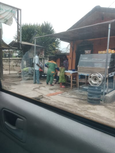 The image captures a street scene in Alagbede, Nigeria, as viewed from inside a vehicle. In the foreground, the interior of a car door is visible, with a dark gray door handle and armrest. The main scene unfolds outside, showing a collection of structures that appear to be market stalls or small shops.

Several individuals are present. A man in a light-colored shirt and blue jeans stands near the center, facing away from the viewer. To his right, a woman in a green jumpsuit with yellow accents stands with her hands on her hips, looking towards the right of the frame. A young girl in a green dress and pink sandals is positioned in front of a wooden table, looking towards a display case. Another woman in a patterned dress is partially visible behind her. A person in a green shirt and gray pants is lying down on a bench or table to the left of the frame.

Objects present include a glass-fronted display case containing various items, likely food. A metal wheel rim sits atop a stack of black tires. Various containers and bags are scattered around the area. A wooden table and chairs are also visible.

The environment appears to be an outdoor, somewhat informal setting. The ground is paved concrete, and there are indications of some wear and tear. In the background, a wall and some vegetation, including spiky-leafed plants, can be seen. A tattered banner with text, including "NA 2025," is attached to a metal pole on the left side of the image. The sky is overcast. The overall impression is a busy, working street scene.