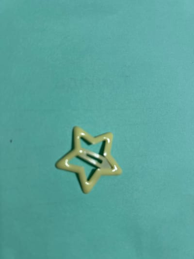 A pale yellow star-shaped hair clip is positioned in the center of the image against a teal background. The clip is made of plastic and has a hollow interior. The background appears to be a flat surface with a slight texture. There are no people or any other objects visible in the image. The location in the city of Дедемель, Kyrgyzstan is not determinable from the provided image.
