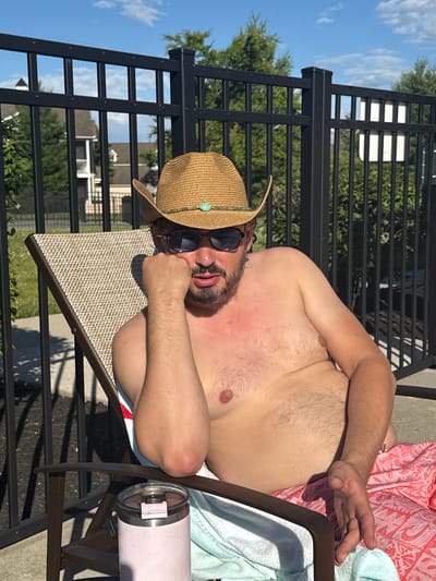 A Caucasian man wearing a straw cowboy hat and sunglasses reclines on a lounge chair by a fence and trees. He is shirtless and wearing patterned shorts. A pink tumbler is visible in the foreground next to a towel. The background shows houses and a blue sky with clouds. The man is resting his head on his hand.