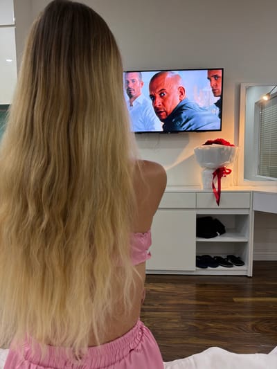 An Hải Ward, Vietnam. An indoor scene features a female individual viewed from behind, her long, wavy blonde hair cascading down her back. She is wearing a two-piece pink garment, consisting of a top revealing her upper back and matching bottoms. Her gaze is directed towards a wall-mounted flat-screen television displaying a movie scene with three male actors, prominently featuring Vin Diesel in the center. Below the television, a white console unit with two drawers on the left supports a large bouquet of red roses, wrapped in white with a red ribbon. The right side of the console includes open shelving, containing folded dark apparel on the upper shelf and two pairs of dark slippers on the lower shelf. To the right, a white vanity table with a mirror reflects a window with horizontal blinds and illuminated string lights. The floor is covered with dark wood planks.
