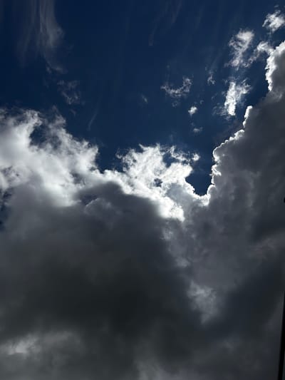 The image depicts a dramatic sky filled with dark, heavy clouds in the foreground, partially obscuring a brighter, sunlit sky with scattered white clouds and wisps of cirrus clouds in the background. The sun is not directly visible but is implied by the bright illumination of the cloud edges and the patches of clear blue sky. The overall impression is one of impending weather change, possibly rain or a storm. The scene is entirely natural, with no human presence or man-made objects prominently featured. The context of Skrzydlna, Poland, suggests this is an outdoor scene taken from ground level, looking upwards.