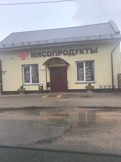 The image shows the exterior of a meat products store in Stowbtsy, Belarus. The building has a light yellow, horizontally sided facade and a gray, corrugated metal roof. A large red logo is on the left side of the building, followed by large, dark letters spelling "МЯСОПРОДУКТЫ" (Meat Products). There are two large windows displaying images of raw meat. A dark red door is located between the windows, under an arched awning decorated with string lights. A sign with information is posted to the right of the door. In front of the store is a sidewalk with a planter on the left and a trash receptacle and bike rack on the right. The sidewalk is wet, indicating recent rain or moisture, with puddles reflecting the building. The street in front of the sidewalk is also wet and appears to be asphalt. A street sign reading "СОВЕТСКАЯ 3" (Sovetskaya 3) is visible on the left side of the building, indicating the street name and building number. The sky is overcast.