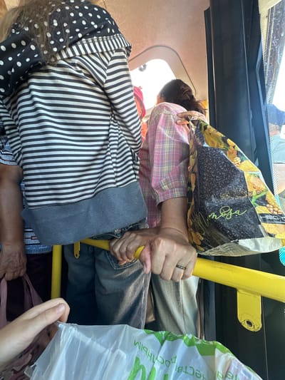 The image depicts an interior view of a crowded bus in Bazhevo, Russia. In the foreground, a person's hands are visible, one holding a yellow metal bar and the other a white plastic bag. To the left, another person's hand holds a pink cloth bag. Behind them, a person wearing a striped shirt with a polka dot hood is seen from the back. Further back, a person in a pink plaid shirt is visible. A large shopping bag with a sunflower print is held by a person on the right. The bus interior appears dimly lit with natural light entering from the windows.
