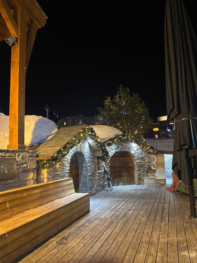 The image depicts an outdoor night scene in the city of Saint-Martin-de-Belleville, France, characterized by a snow-covered, alpine-style environment. The foreground features a wide wooden plank deck. To the left, a multi-tiered wooden bench is oriented towards the center of the frame, with a large wooden structural pillar partially visible on the far left edge. To the right, a tall, dark, folded outdoor umbrella or canopy stands upright.
In the midground, a rustic stone building with a slate roof is prominent. The building features two distinct arched doorways, each fitted with closed wooden doors. The roof is partially covered with snow, and both the roofline and the arches are decorated with green garlands and illuminated string lights. A pair of skis and ski poles lean against the stone wall between the two arched entrances. Behind this structure, a large evergreen tree adorned with string lights is visible.
The background reveals several illuminated buildings, indicative of a village or resort, with additional snow accumulation on roofs and ground. Faint outlines of ski lift components are discernible against the dark night sky. No individuals are present in the scene. The artificial lighting from the decorations and background structures illuminates the immediate area.