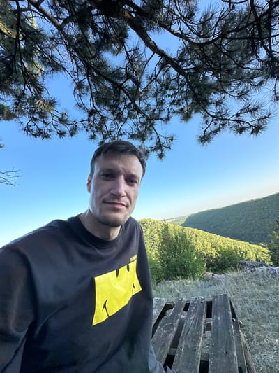 A man in a black t-shirt with a yellow graphic on it is taking a selfie outdoors. He is framed from the chest up. Behind him is a lush green valley with rolling hills, and trees. The sky is a clear, bright blue. The man appears to be standing on a wooden structure that looks like a picnic table or platform. Pine branches hang over the top of the image, partially obscuring the view of the sky. The location is the city of Pryvorottia Druhe, Ukraine.