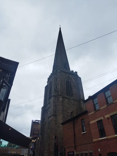 The image captures St Peter's Church on St Peter's Street in Hereford, United Kingdom, under an overcast sky. The central feature is the church's imposing grey stone tower, topped with a tall, dark, four-sided spire culminating in a finial. The tower exhibits pointed arched windows with louvers and decorative battlements below the spire's base. To the left, a dark building with large, multi-paned windows is partially visible, beneath which a dark awning extends over a lower area. Text reading "GABI" and "POLISH SHOP" is partially visible below the awning. Adjacent to the church tower, a dark rectangular sign for "Behind The Wall Freehouse" hangs prominently. Below this sign, a blue circular road sign is partially obscured. To the right of the church, a multi-story building constructed of red brick with cream-colored banding and dark-framed windows is visible. The ground level of this building displays partial lettering for "CARDZONE". Several dark utility lines traverse the grey, cloudy sky. No individuals or specific actions are discernible within the frame.