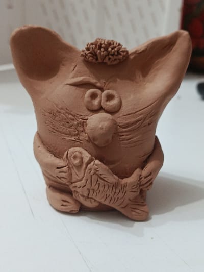 A close-up, eye-level, medium shot captures a small, unglazed clay figurine of a whimsical creature holding a fish. The creature has large, cartoonish eyes, prominent ears that extend outwards, and a small tuft of textured clay resembling hair on its head. Its body is somewhat squat and rounded, with sculpted lines suggesting a mouth and perhaps a snout. The creature's arms are wrapped around a stylized fish, which is depicted with scales and fins. The figurine is made of a reddish-brown clay. The background is blurred and out of focus, showing a light-colored, possibly white, surface that appears to be a tabletop with faint lines, and a hint of a reddish-brown object on the right side. The lighting is soft, creating subtle shadows. The scene is indoors, and there is no indication of its location within the city of Ashgabat, Turkmenistan, or any other specific geographic location.