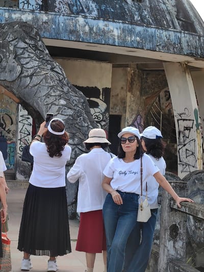 The image depicts five women standing outdoors in Kim Long Ward, Vietnam, in front of a large, weathered concrete structure. This structure, part of the Thien Mu Pagoda's entrance area, features a prominent grey, scaled, dragon-like architectural element on its left side. The concrete surfaces exhibit significant weathering, blue-green discoloration, and extensive graffiti tags in various colors, including black, white, and red, with a distinct "26" in a white circle visible on one wall.
In the foreground, a woman wearing a white short-sleeved t-shirt with "Girly Things" text, blue jeans, a white baseball cap, and dark sunglasses poses for the camera. Her left hand rests on a weathered concrete railing. Partially obscured behind her is another woman wearing a white cap and top. To the left, a woman seen from the back wears a white visor, a white short-sleeved top, and a long black pleated skirt. She holds a white smartphone vertically, appearing to photograph the scene. Beside her, another woman, also facing away, wears a beige bucket hat, a white long-sleeved shirt, and a reddish-brown knee-length skirt. A fifth woman is partially visible on the far left. The background reveals an interior concrete staircase within the structure, also covered in graffiti. The scene is brightly illuminated by daylight.
