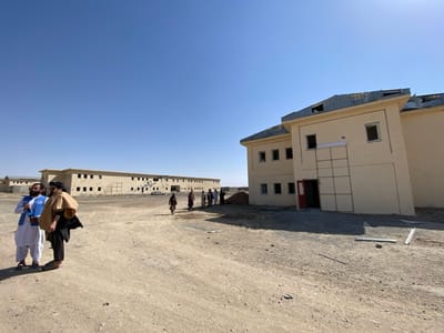 The image depicts an arid outdoor environment in Bar Mel, Afghanistan, under a clear blue sky. The ground is dry, uneven dirt and gravel, with scattered debris near the foreground structure. In the immediate foreground, two men stand. The man on the left wears a white shalwar kameez, a dark vest, and a light turban, holding a blue object. The man on the right wears dark clothing with a tan cloak draped over his shoulders and a dark turban. To the right, a two-story cream-colored building with a partially damaged corrugated metal roof is visible. The roof shows lifted and possibly missing sections. The building's facade features multiple rectangular window openings, some lacking glass, and a red entrance door. A large, grid-patterned panel is affixed to the wall adjacent to the entrance. In the mid-ground, stretching horizontally, is a longer, lighter-colored building, also with a corrugated metal roof and numerous rectangular openings, some exhibiting dark marks consistent with damage. A group of approximately six individuals, dressed in dark traditional attire, walks on the dirt path between the buildings, moving away from the viewer towards the longer structure.