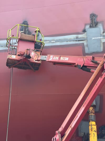 In Geoje-si, South Korea, two workers, clad in safety vests and hard hats, are positioned in the basket of a cherry picker. The cherry picker is extended upwards, reaching a large, red, curved surface, likely the hull of a ship under construction or repair. The surface exhibits areas of gray primer and dark, possibly burnt, patches, suggesting recent welding or repair work. One worker appears to be actively engaged in a task, while the other observes. The overall environment suggests an industrial shipyard setting.