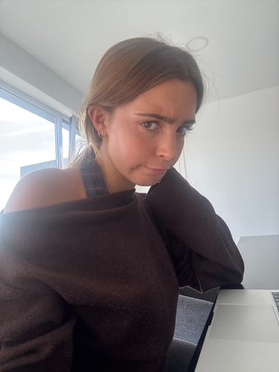 A close-up image captures a young woman looking directly towards the viewer with a calm, pensive expression, her head slightly tilted. She has light brown hair, some wisps around her forehead, and brown eyes. A small, dark mark is visible on her right cheek. She is wearing a dark brown, textured garment, possibly a robe or blanket, draped over her shoulders and right arm. A subtle plaid strap is visible underneath this garment. A delicate, cross-shaped earring is worn in her right ear.
The scene is indoors, brightly lit by natural daylight streaming through a window visible in the background to her left. The window frame is present, and the wall or ceiling above her is light-colored. To her right, a silver laptop, likely a MacBook, with its trackpad visible, rests on a dark surface next to a dark grey upholstered chair or sofa. The presence of the laptop suggests a relaxed, everyday setting, possibly a home in Torres Vedras, Portugal. The time of day is clearly daytime due to the bright natural light. No visible text is present in the image.