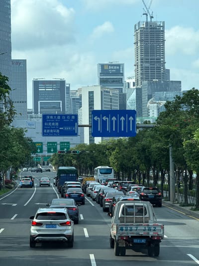 The image captures a traffic scene in Nanshan District, Shenzhen, China. A multi-lane road is densely packed with cars and trucks moving forward. A blue overhead traffic sign indicating lane directions is prominently visible, along with street signs for Shennan Blvd, Baishi Rd, Keyuan South Rd, and Gaoxin South Ring Rd. Tall buildings and skyscrapers are observable in the background, including a building under construction and several bearing business logos such as "CR LAND". Trees line the road alongside the traffic. The traffic lights are green. Vehicle license plates are visible, with designations such as "B-83750" and "B-87JW2".