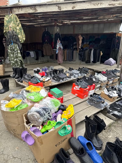 The image shows a market stall in Ust-Kan, Russia. Clothing hangs from racks on the left side of the stall. Shoes, including sneakers, boots, and colorful Crocs-style clogs, are displayed on wooden platforms and in a cardboard box on the right. Some shoes are in boxes, some are in plastic bags. The box on the right has “CCCA” in Russian on the side.