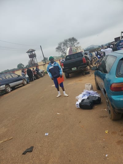 The image depicts an outdoor scene in what appears to be Ohit, Nigeria. In the foreground, a young male wearing a green beanie, a blue and white track jacket, blue shorts, and white sneakers stands holding an orange folder. To his right, a pile of white folded clothing and black plastic bags are on the ground next to a teal car. In the background, several people are gathered near parked vehicles, including a black pickup truck. A tall observation tower with a metal structure and a small roofed enclosure at the top is visible to the left. The ground is paved with reddish-brown dirt and asphalt. The sky is overcast, suggesting cloudy weather.