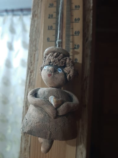A close-up, eye-level shot captures a handmade clay figurine of a girl with light blue eyes and red lips, wearing a brown textured hat and a dress. She is suspended by a thin string in front of a wooden thermometer. The thermometer has markings from 0 to 80, with numbers visible at 0, 10, 20, 30, 40, 60, and 80. The figurine is positioned slightly to the left of the thermometer. The background consists of a blurred white curtain or fabric on the left and a dark wooden surface on the right. The lighting is subdued, highlighting the textured surface of the clay figurine. The scene appears to be an indoor setting, possibly a home or a workshop in the city of Shelekhov, Russia, given the context of the thermometer often used for weather monitoring.