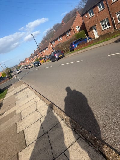The image presents a residential street scene in Rotherham, United Kingdom, under bright, clear conditions with a blue sky and scattered white cumulus clouds. The perspective is from the pavement, looking down a gently curving road lined with houses.
In the immediate foreground, the elongated shadows of two individuals are cast diagonally across the light grey concrete slab pavement and extend onto the dark asphalt road, indicating a low sun angle. The pavement exhibits minor cracks and variations in tone. A textured, granular curb separates the pavement from the roadway. The road surface is dark grey asphalt, marked with white dashed lane lines centrally and a solid white line along the curb.
A row of two-story, red brick residential houses with brown tiled roofs, white-framed windows, and white front doors lines the right side of the street. These properties feature small front gardens with hedges and grass verges. A dark grey/purple Nissan Micra (license plate YAG5 OTY) is parked on the right, partially on the road. Further along the street, a blue Skoda Octavia estate is parked partially on a verge. Additional vehicles, including a black hatchback and a white sedan, are visible further down the road, some parked and others potentially in motion.
Black, pole-mounted streetlights are spaced along the road. Overhead power lines are visible against the sky. In the background, a dense line of deciduous trees with bare branches borders the residential area.