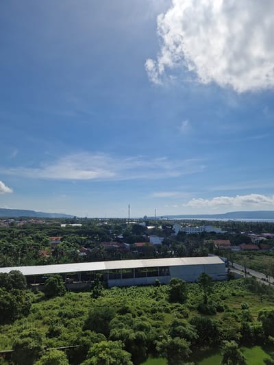 The image shows a view of Banyuwangi, Indonesia, from an elevated position. The sky occupies the left portion of the frame with scattered clouds. In the center, a dense residential area transitions from the buildings near the coast to a mountainous region in the distance. A prominent gray building runs vertically through the right side of the frame, surrounded by green foliage. A road runs parallel to the building near the top of the frame. The photograph seems to capture a clear, sunny day. There are no people visible in the image.