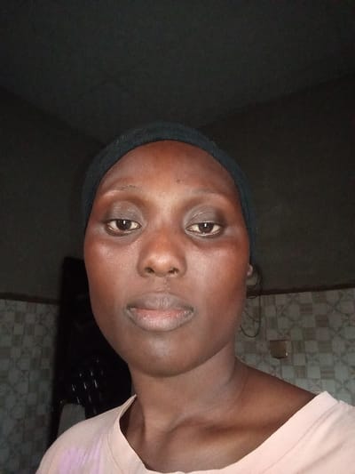 A close-up, chest-up portrait features a person with dark skin, likely a woman, looking directly into the camera with a neutral expression. She wears a dark headband or head covering that pulls her hair back from her forehead, and a light-colored, possibly pale pink or beige, t-shirt.
The background indicates an indoor setting. On the lower left and right, a patterned wall is visible, possibly with tiles or textured wallpaper in earthy tones. The upper background and ceiling are dark and indistinct. The lighting appears to be frontal, illuminating the subject's face, which shows some natural skin texture and subtle highlights. There are hints of slight puffiness or darkness under the eyes.
The subject is posing for the photograph; no other activity is depicted. The indoor lighting does not provide cues for the time of day or weather. No visible text is present in the image. The setting is a room in Fanisau, Nigeria.