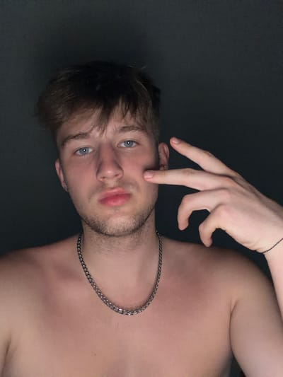 A young Caucasian male with light brown hair and blue eyes is captured in a close-up, chest-up shot against a dark, nondescript background. He is shirtless, wearing a silver chain necklace. His left hand is positioned near his face, with three fingers extended and touching his cheek. A dark string bracelet is visible on his right wrist. The lighting appears to be from the front, casting subtle shadows. The image does not contain any discernible features that would indicate a specific location in Vanino, Russia.